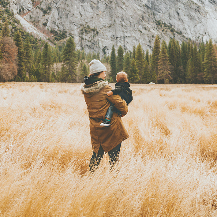 Mother and child stand in a meadow with trees and a mountain behind